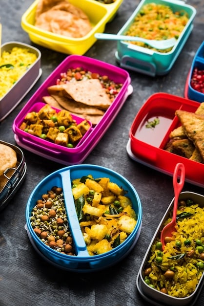 A close-up shot of several neatly organized, labeled meal prep containers filled with colorful and healthy food, such as grilled chicken, roasted vegetables, and quinoa. Hands are seen in the background, possibly packing or arranging the containers.