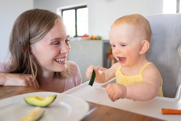 A mother smiling and watching her baby confidently grasp and eat a soft slice of avocado during a baby-led weaning meal.