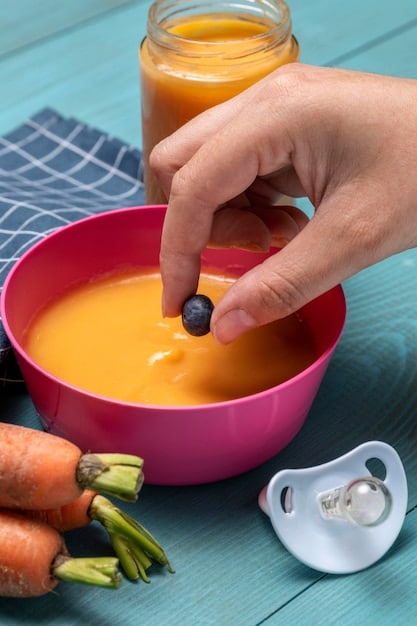 A close-up shot of a mother feeding her 6-month-old baby pureed carrots with a small spoon. The baby is looking at the mother with a curious and happy expression, ready to take another bite. The background is a cozy and bright dining area with natural light.