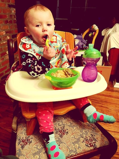 A baby sitting in a high chair, happily reaching for a spoon filled with vibrant orange sweet potato puree. The baby's face is slightly messy but filled with joy and curiosity, with the mother's hand gently guiding the spoon.