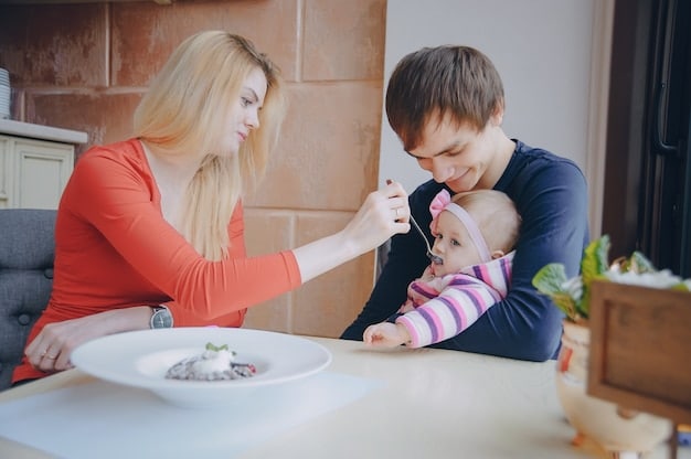 A family (mother, father, and baby) are sitting around a table. The baby is in a high chair, reaching for small pieces of soft vegetables from their plate. The parents are smiling and encouraging the baby's self-feeding efforts.