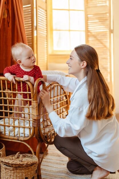 A mother gently supports her baby in a cradle hold, carefully watching the baby's face. The baby's eyes are open and alert, and the baby's mouth is slightly agape as if ready to feed. The lighting is soft, highlighting the gentle interaction between mother and child.