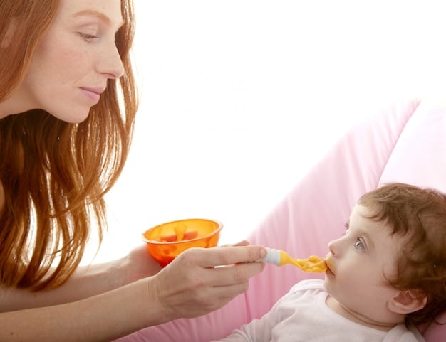 A mother feeding her baby mashed sweet potatoes from a spoon. The baby is looking at the mother with a happy expression.
