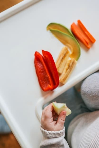 A close-up shot of a baby's hands exploring a small pile of soft, colorful vegetable sticks (carrots, cucumbers, bell peppers) on a high chair tray. The baby is actively touching and examining the different textures and shapes, with a look of curiosity and concentration. The background is blurred to keep the focus on the baby's hands and the food.