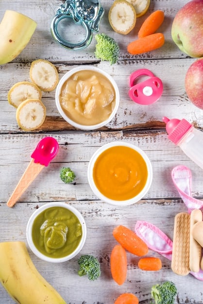 A diverse spread of colorful, baby-friendly solid foods, including pureed fruits, steamed vegetables, and soft finger foods, arranged on a clean, child-friendly surface.
