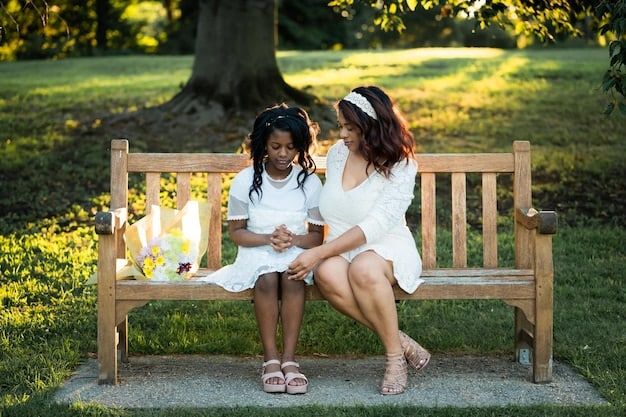 Two moms sitting on a park bench, facing each other and smiling. One mom is holding a baby in a carrier, and the other is reaching out to touch the baby's hand gently. The background is blurred with greenery.