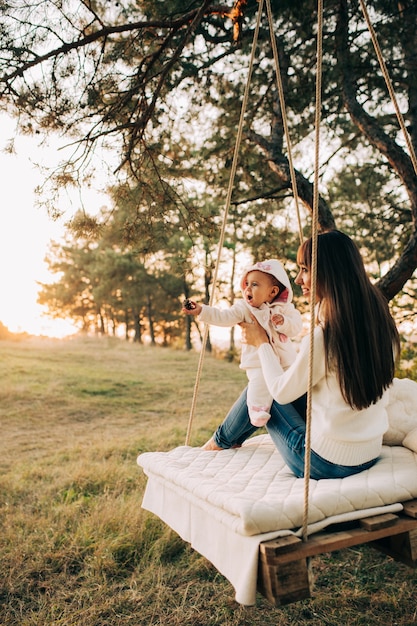 A mom smiling as she talks on the phone, sitting on a park bench. Her child is playing happily on the swings nearby. The sun is shining, and the overall mood is light and joyful.