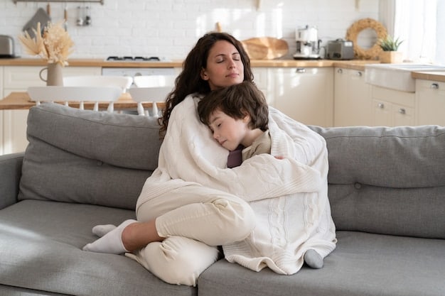 A young mother is peacefully napping on a couch. Her baby lies asleep on her chest, and a plush blanket covers them both. Sunlight streams through the window, creating a cozy and serene atmosphere.