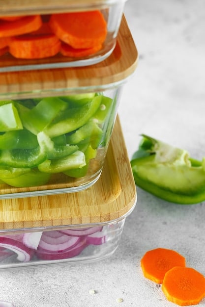 A close-up of various pre-cut vegetables (carrots, celery, bell peppers) being stored in clear, airtight containers in a refrigerator. Labels on the containers indicate the date and contents.