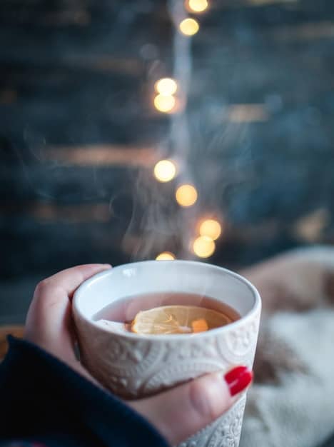 A close-up of a woman's hands holding a warm cup of herbal tea by candlelight, with a background of soft, blurred fairy lights.