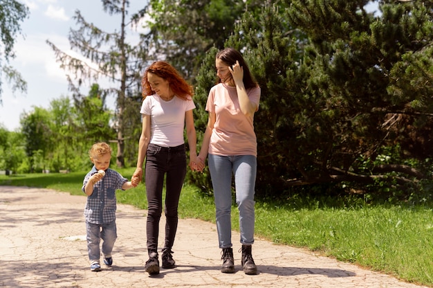 Three moms are walking down a suburban street together, pushing strollers. They are talking and smiling at each other. The houses and lawns are well-kept.