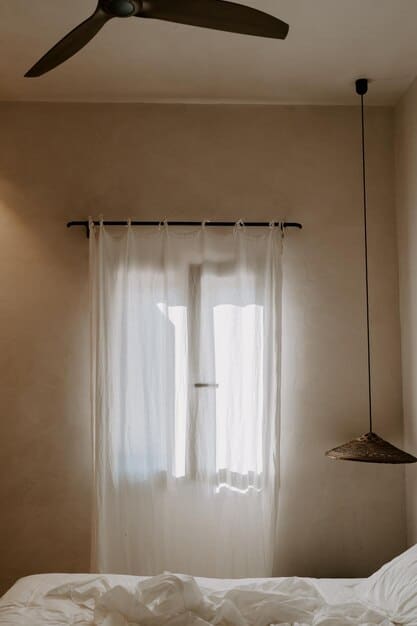 A bedroom with an open window, sheer curtains blowing gently in the breeze, and a small fan placed on a nightstand. The room is decorated in calming, neutral tones.